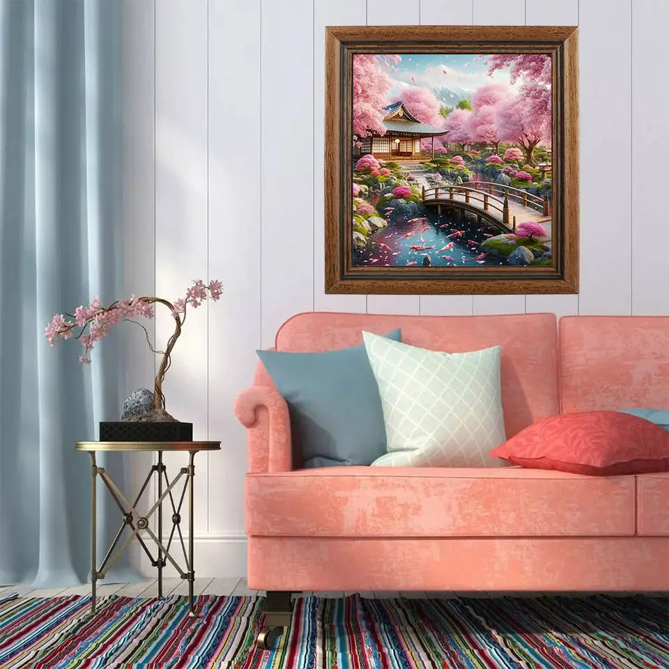 Beautiful bright pink deluxe 5D diamond painting kit featuring a traditional Japanese house, flowers, bridge and a pond full of bright swimming koi fish hung in a beautiful  brown frame hung up in  a modern living room above a matching pink couch. 