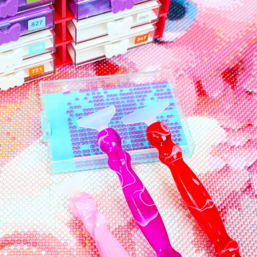 Colorful diamond painting tools on a pink textured surface with books in the background